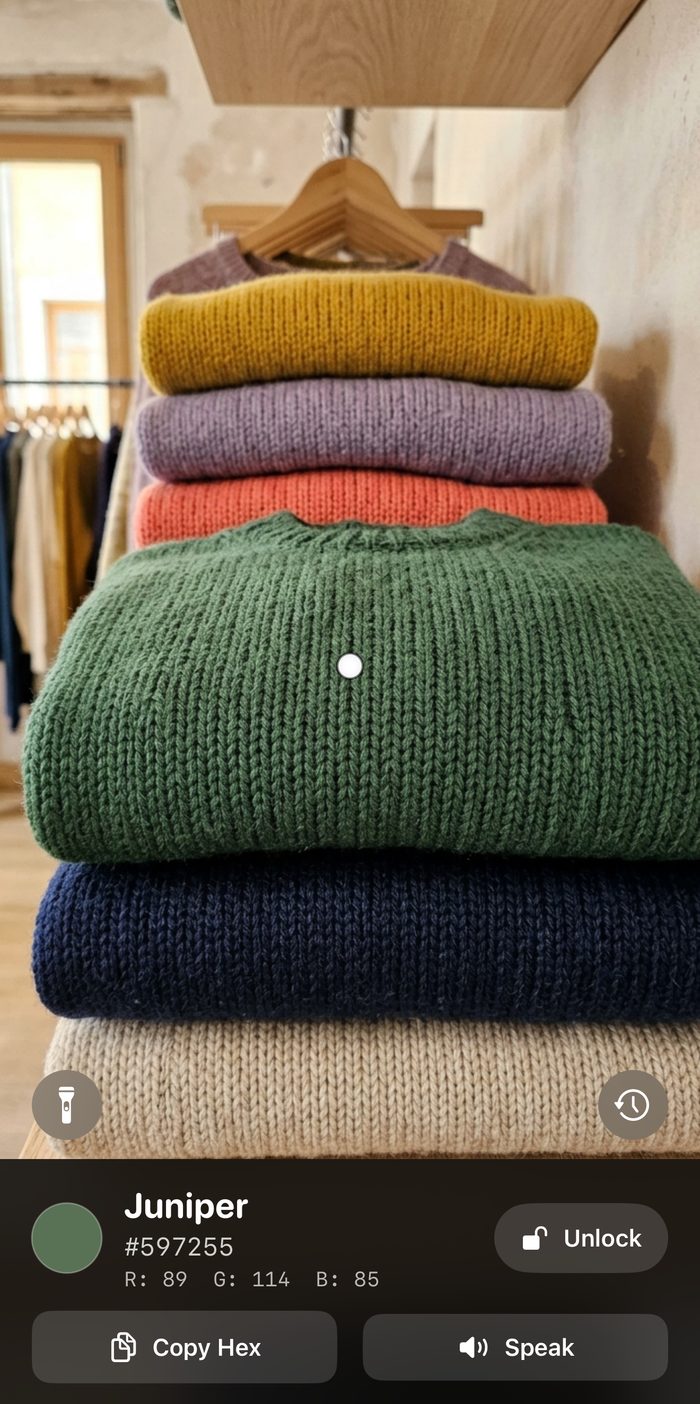 Hue pointed at a stack of knitted sweaters, locked on the green one. The panel reads Juniper, hex 597255, red 89, green 114, blue 85, with Copy Hex and Speak buttons.